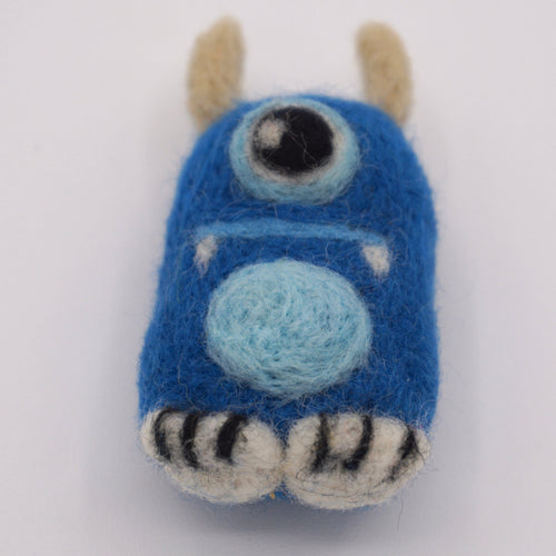 A small felted wool monster toy in bright blue, with a light blue circular patch, black and white striped feet, beige horns, and white accents, photographed on a plain white background.